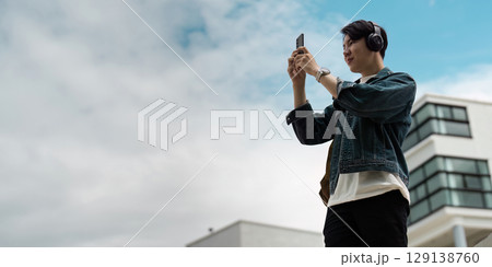 Young Man Enjoying Music and Photography Outdoors with Smartphone and Headphones Against Modern Urban Background 129138760