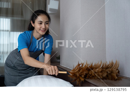 Woman Dusting a Modern Bedroom with Feather Duster in a Cozy Home Environment 129138795