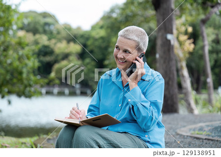 Senior Woman Enjoying Outdoor Lifestyle While Engaging in a Phone Call and Taking Notes in a Serene Park Setting Senior Woman Enjoying Outdoor Lifestyle While Engaging in a Phone Call and Taking Notes in a Serene Park Setting 129138951