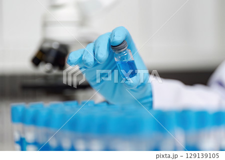 Scientist Holding Blue Liquid in Laboratory Vial with Microscope in Background for Scientific Research and Experimentation 129139105
