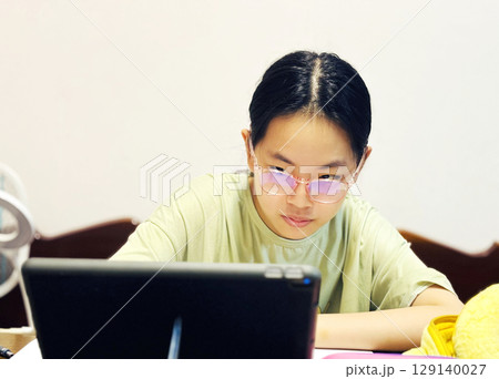Portrait of an Asian preteen, 12-13 years old. The child is studying lessons online at home, wearing protective eyeglasses, a real-situation image of an Asian homeschool 129140027