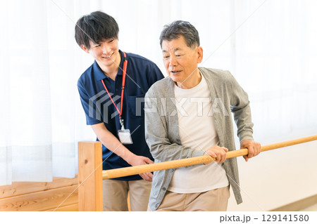 手すりを使い歩行訓練するシニア男性と介助する作業療法士,理学療法士の男性スタッフ 手すりを使い歩行訓練するシニア男性と介助する作業療法士,理学療法士の男性スタッフ 129141580