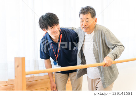 手すりを使い歩行訓練するシニア男性と介助する作業療法士,理学療法士の男性スタッフ 129141581