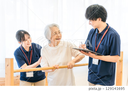 手すりを使い歩行訓練するシニア女性と介助する作業療法士,理学療法士の男女スタッフ 129141695