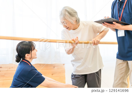 手すりを使い歩行訓練するシニア女性と介助する作業療法士,理学療法士の男女スタッフ 129141696