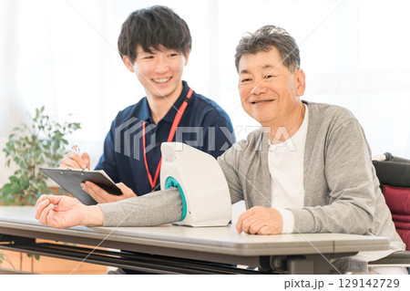 血圧計を使い血圧測定をするシニア男性と記録をとる看護師,介護士の男性スタッフ 血圧計を使い血圧測定をするシニア男性と記録をとる看護師,介護士の男性スタッフ 129142729