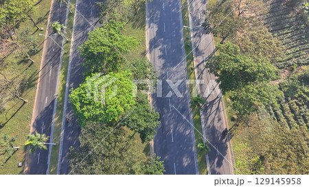 An aerial view of a wide, multi-lane road flanked by trees, with cars and motorbikes driving on a sunny day. An aerial view of a wide, multi-lane road flanked by trees, with cars and motorbikes driving on a sunny day. 129145958