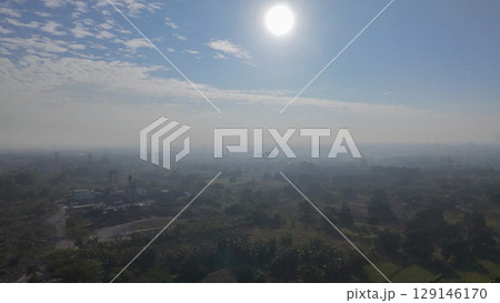 A hazy, sun-drenched aerial view of a sprawling landscape, with a mix of fields, trees, and scattered buildings under a bright sky with wispy clouds. A hazy, sun-drenched aerial view of a sprawling landscape, with a mix of fields, trees, and scattered buildings under a bright sky with wispy clouds. 129146170