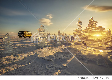 Sunset reflected in a large block of ice hummock and a classic vintage van car on the frozen lake background. 129147249