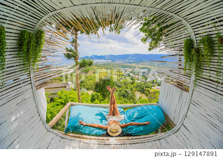 Beautiful slim tourist woman is lying down sunbathing, covered had with huge straw hat, near terrace in cafe outdoor with scenic view on mountains. Time to relax. 129147891