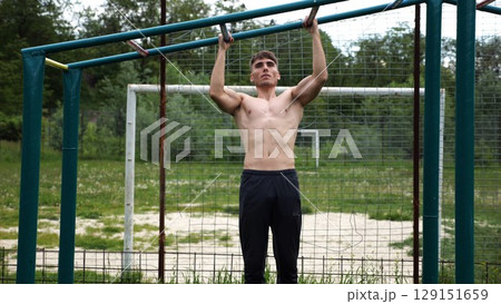 Young muscular sportsman doing pull ups at sports ground. Athletic guy exercising outdoor. Strong sporty man working out at summer nature. Concept of healthy and active lifestyle. Slow motion 129151659