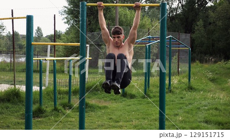 Strong guy training abdominal muscles on horizontal bar at sports ground. Young sportsman exercising on pull-up bar outdoor. Sporty athlete working out at nature. Concept of sportive lifestyle Strong guy training abdominal muscles on horizontal bar at sports ground. Young sportsman exercising on pull-up bar outdoor. Sporty athlete working out at nature. Concept of sportive lifestyle 129151715