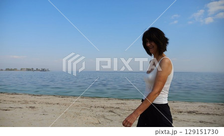 Happy adult woman going on sandy beach near shoreline at sunny day. Beautiful lady walking on seashore with calm waves at background. Concept of summer vacation or holiday. Slow motion 129151730