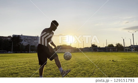 Young man juggling soccer ball on stadium at sunset. Professional footballer kicking ball at green field. Sportsman practicing tricks at meadow with sunlight at background. Freestyle football Young man juggling soccer ball on stadium at sunset. Professional footballer kicking ball at green field. Sportsman practicing tricks at meadow with sunlight at background. Freestyle football 129151793