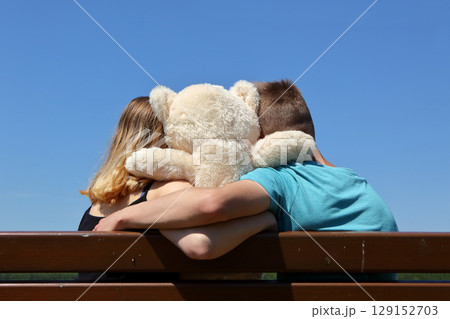 A lovely couple enjoying the company of a cute teddy bear while sitting on a park bench A lovely couple enjoying the company of a cute teddy bear while sitting on a park bench 129152703