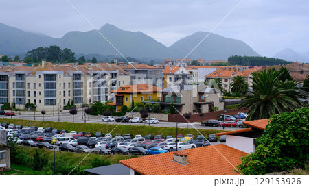 Cityscape of a Llanes, Spain with a mix of modern and traditional buildings, a crowded parking lot, and majestic mountains in the background under a cloudy sky 129153926