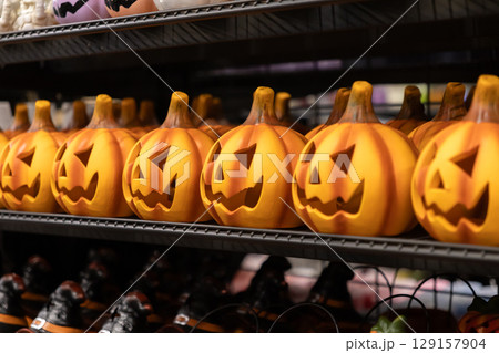 Orange Halloween pumpkin decorations with spooky carved faces are neatly displayed on a store shelf, symbolizing festive seasonal sales and autumn celebration Orange Halloween pumpkin decorations with spooky carved faces are neatly displayed on a store shelf, symbolizing festive seasonal sales and autumn celebration 129157904