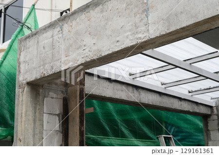 Concrete structure framing with exposed metal beams supporting translucent sheet roof. Green netting covers building wall area. Construction site showing progress. Materials visible Concrete structure framing with exposed metal beams supporting translucent sheet roof. Green netting covers building wall area. Construction site showing progress. Materials visible 129161361