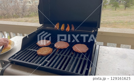 View of an open gas grill with colorful carrots and veggie patties cooking, with a hand reaching for buns on the side. Great for showcasing outdoor cooking setups or plant-based grilling ideas. View of an open gas grill with colorful carrots and veggie patties cooking, with a hand reaching for buns on the side. Great for showcasing outdoor cooking setups or plant-based grilling ideas. 129161625