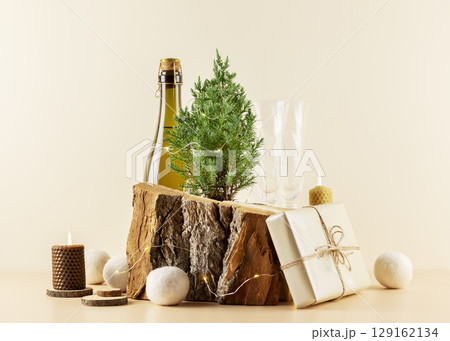 Winter holiday composition with bottle of champagne, glasses, small fir tree, gift and candles on a wooden podium on a beige background. Christmas and New Year Card 129162134