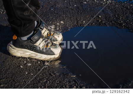 Close-up of children's feet in sneakers standing on a dirt road near a puddle 129162342