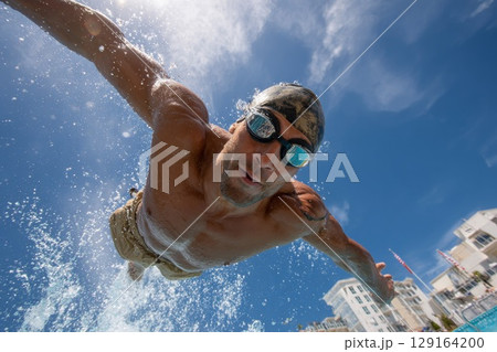 Male diver performing a dynamic springboard dive into a clear blue swimming pool 129164200