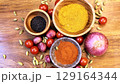 Colorful spices nestled in wooden bowls, scattered with fresh vegetables and aromatic pods, presenting rustic culinary ingredients on weathered wooden surface 129164344