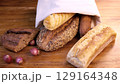 Artisan bread varieties spilling from crumpled white bag, nestled on weathered wooden surface alongside rustic onions, embodying traditional bakery warmth 129164348