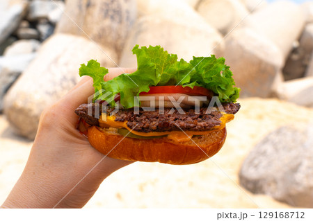 Woman holding burger in a hand against beach background 129168712