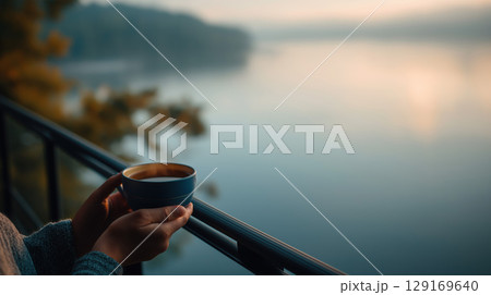 Person holding cup, peaceful morning, lake view, soft light, outdoor relaxation, tranquil mood 129169640