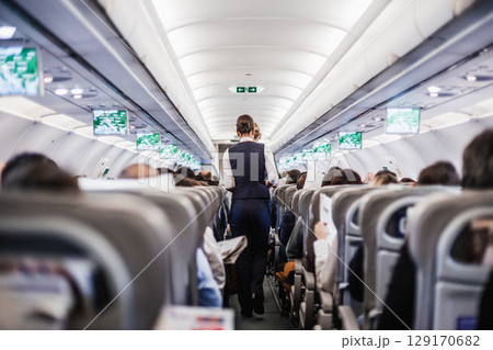Interior of airplane with passengers on seats and stewardess in uniform walking the aisle, serving people. Commercial economy flight service concept 129170682