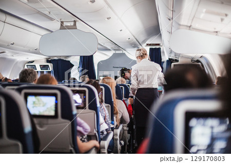 Interior of airplane with passengers on seats and stewardess in uniform walking the aisle, serving people. Commercial economy flight service concept 129170803