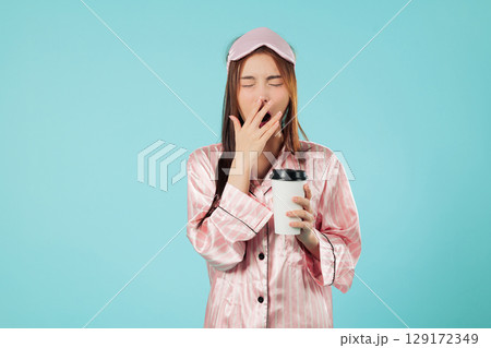 Tired young woman in pajamas and sleep mask holding coffee cup, needs that morning coffee to wake up as she rubs her eye while resting at home, headshots isolated studio blue background 129172349