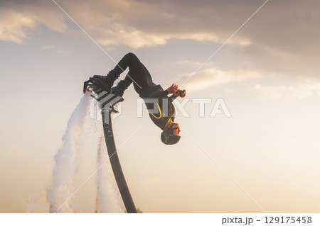 Athlete flyboarder doing close-up stunts. Exciting flyboarding experience at sunset showcasing impressive aerial maneuvers above sparkling water surface Athlete flyboarder doing close-up stunts. Exciting flyboarding experience at sunset showcasing impressive aerial maneuvers above sparkling water surface 129175458