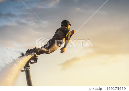 A flyboarder displays skill and excitement while soaring above the water during a stunning sunset. The bright colors of the sky create a dramatic backdrop as the rider navigates through the air. A flyboarder displays skill and excitement while soaring above the water during a stunning sunset. The bright colors of the sky create a dramatic backdrop as the rider navigates through the air. 129175459