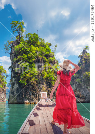 A Woman female solo traveler in a bright red dress and a wide-brimmed straw hat Standing on a Traditional Thai Longtail Boat and looking at the broken rocks on Cheo Lan Lake in Khao Sok Park in 129175464