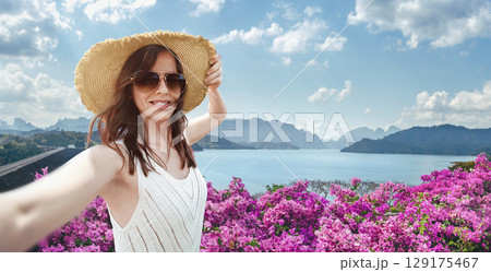 European woman travel blogger in a straw hat and sunglasses takes a selfie photo at the observation point Rachaphrapha dam Cheo Lan Lake in Khao Sok Park in Thailand. Solo travel in Southeast Asia 129175467