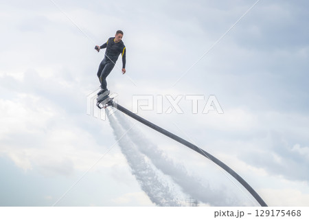 A man in a black athlete wetsuit hovers against a dramatic sky on a flyboard. A performer demonstrates impressive flyboarding skills, soaring above the clear blue water. Dressed in a sleek wetsuit, 129175468