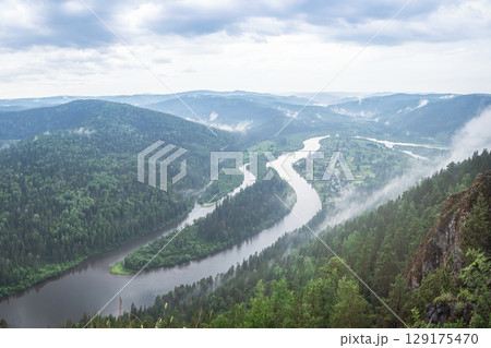 Scenic view of a winding river cutting through lush green hills in Siberia, surrounded by mist and clouds 129175470