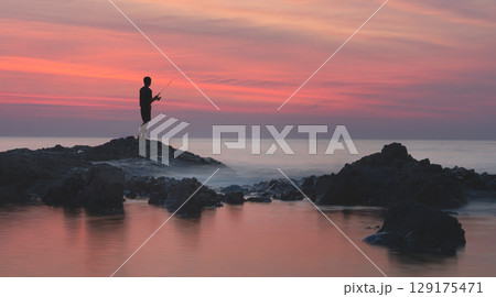 silhouette of a male fisherman above the foam waves. He is spinning fishing oastal stoneridge in Khao Lak. water is filled with red glare shining sunset. Silhouette man against the backdrop of a silhouette of a male fisherman above the foam waves. He is spinning fishing oastal stoneridge in Khao Lak. water is filled with red glare shining sunset. Silhouette man against the backdrop of a 129175471