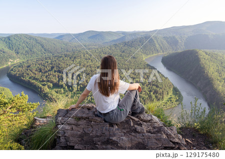 A person sits on a rocky ledge, gazing at a breathtaking view of a winding river framed by rolling hills and dense forests. A female solo traveler rests on a mountain and looks at the Mana River. The A person sits on a rocky ledge, gazing at a breathtaking view of a winding river framed by rolling hills and dense forests. A female solo traveler rests on a mountain and looks at the Mana River. The 129175480