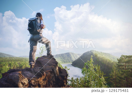 Hiker standing on rocky ledge overlooking river and forest landscape during daytime under blue sky . A female traveler with a backpack on the Mana River and the Manskaya Loop in Siberia, Russia Hiker standing on rocky ledge overlooking river and forest landscape during daytime under blue sky . A female traveler with a backpack on the Mana River and the Manskaya Loop in Siberia, Russia 129175481