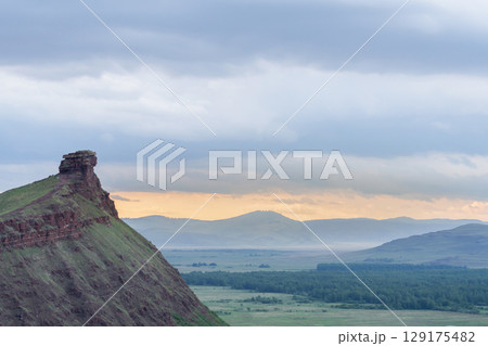 Majestic rock formation overlooks expansive green valley and distant mountains at sunset in a serene landscape. The Trunks mountain range in Khakassia, Russia. Majestic rock formation overlooks expansive green valley and distant mountains at sunset in a serene landscape. The Trunks mountain range in Khakassia, Russia. 129175482