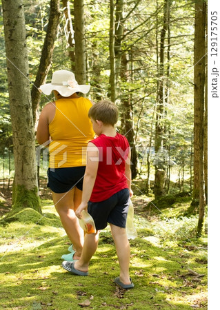 A woman and child hike through a sun-dappled forest, carrying empty water bottles. Enjoying nature's beauty together. 129175705