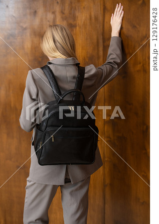A stylish young woman, long blonde hair, poses in a grey suit, holding a brown leather backpack against a warm wooden background A stylish young woman, long blonde hair, poses in a grey suit, holding a brown leather backpack against a warm wooden background 129176428