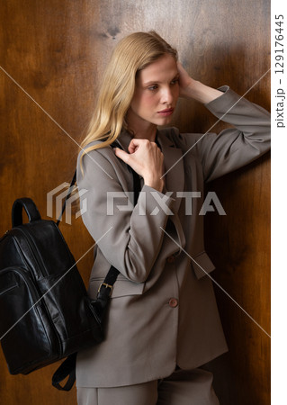 A stylish young woman, long blonde hair, poses in a grey suit, holding a brown leather backpack against a warm wooden background A stylish young woman, long blonde hair, poses in a grey suit, holding a brown leather backpack against a warm wooden background 129176445