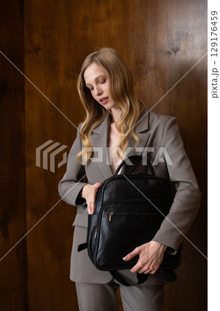 A stylish young woman, long blonde hair, poses in a grey suit, holding a brown leather backpack against a warm wooden background A stylish young woman, long blonde hair, poses in a grey suit, holding a brown leather backpack against a warm wooden background 129176459