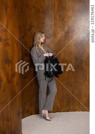 A stylish young woman, long blonde hair, poses in a grey suit, holding a black leather backpack against a warm wooden background 129176461