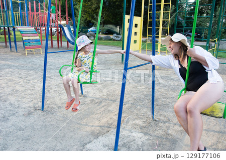 A woman gently pushes a girl on a swing at a playground. They share a sweet moment of playful interaction. 129177106