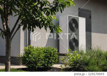 Modern outdoor heat pump unit installed near a residential house wall, surrounded by green shrubs and a tree in the landscaped yard 129178943